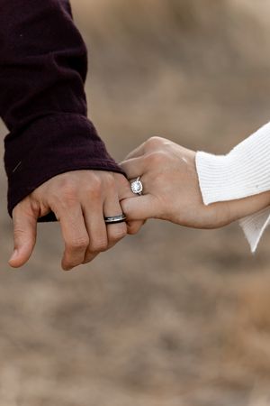 Close-up of couple holding hands, woman wearing diamond engagement ring, man wearing black wedding band.