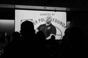 Silhouetted audience members watch a projected image of a bald, bearded man in a dark polo at a donation event in black and white.
