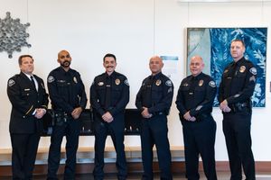 Six uniformed police officers stand side by side in a bright hallway, posing for a group photo in front of blue abstract artwork.