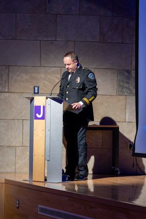 A uniformed police officer speaks at a wooden podium with a microphone during an event in a stone-walled room.