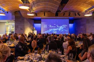 Guests seated at round banquet tables listen to a speaker at a podium during an evening awards dinner beneath a large projection screen.