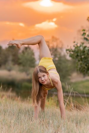 Flexible young gymnast performs a one-handed cartwheel in yellow outfit against golden sunset sky.