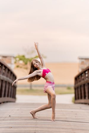 Young dancer in pink outfit performs graceful pose on wooden bridge outdoors.
