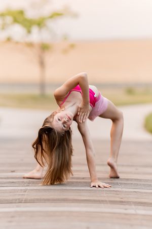 Young girl in pink sportswear performing a backbend pose on wooden deck outdoors.