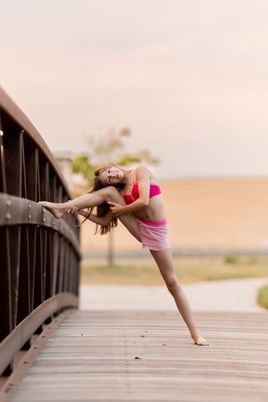 Young flexible dancer stretching on a wooden bridge at sunset, wearing a pink sports top and shorts, smiling gracefully.