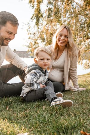 A family sits together on grass during golden hour under autumn trees beside a lake.