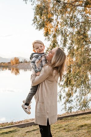 Parent and child share a playful moment by a lake during autumn with golden leaves on trees in background.