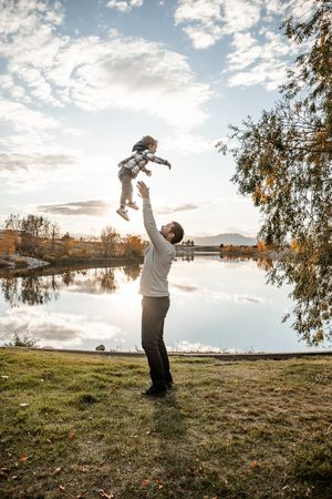 A person lifts a small child into the air against a scenic lakeside backdrop with cloudy skies at sunset.