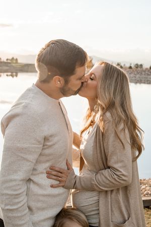 A romantic couple in neutral sweaters sharing tender kisses by the waterfront at sunset.