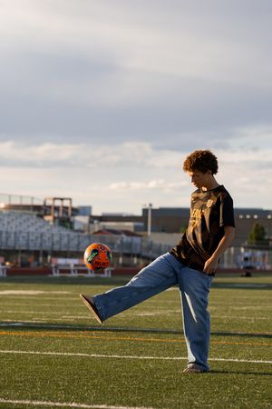 Teen boy in jeans and black jersey kicking a soccer ball on a green turf field under cloudy skies.