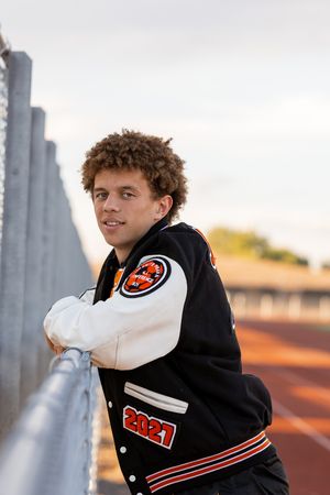 Young man in a 2021 letterman jacket leaning on a railing at a track, wearing curly hair.