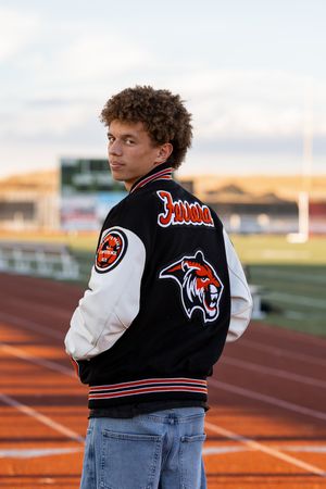 Young man in varsity letterman jacket poses on a track field at dusk, looking over his shoulder.