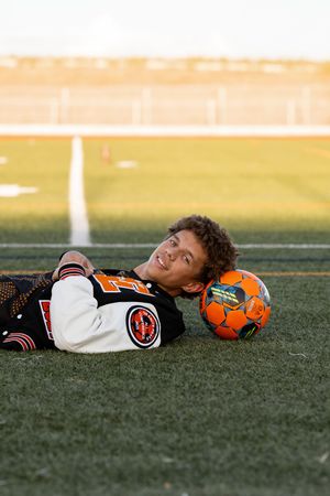 Teen boy in varsity jacket lying on soccer field with orange ball, smiling at camera during golden hour.