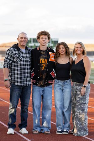Family of four smiling together on a high school track, teen boy wearing a varsity letterman jacket numbered 7.