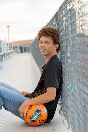 Teen boy with curly hair sits against chain-link fence holding orange soccer ball, wearing black shirt and jeans.