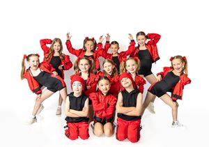 Young girls in red and black dance costumes pose energetically for a group photo against a white background.