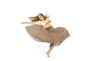 A barefoot dancer in a flowing tan skirt and cream top leaps gracefully against a white background.
