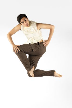 Barefoot male dancer mid-jump in white sleeveless top and brown pants, performing dynamic contemporary dance move.