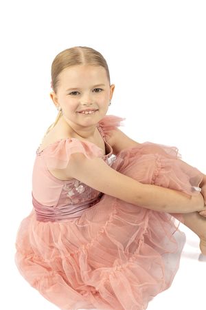 A young girl in a pink tulle dress sits smiling on a white background, wearing earrings and looking elegant.