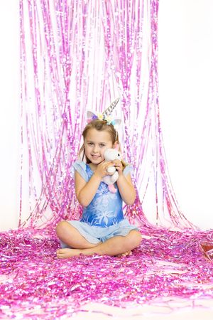Child in blue dress sitting on pink sparkly backdrop wearing unicorn horn and holding stuffed animal.