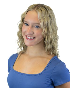 A person with wavy blonde hair in a royal blue top smiles while posing against a white background.