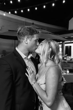 A romantic couple shares a tender kiss in a dimly lit venue with string lights overhead.