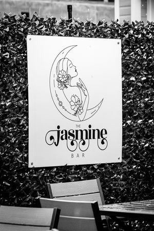 Sequence of black and white business signs with floral designs and 'Jasmine' text mounted on outdoor wall with leafy backdrop.