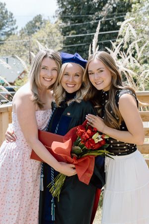 Three women smile together outdoors, the center one wearing a graduation cap and gown holding red flowers.