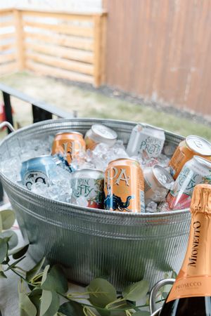Galvanized metal tub filled with ice, assorted beer cans, and a Mionetto Prosecco bottle at an outdoor party.