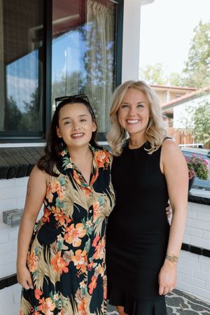 Two smiling women posing together outdoors, one in a floral dress and one in a black sleeveless dress.