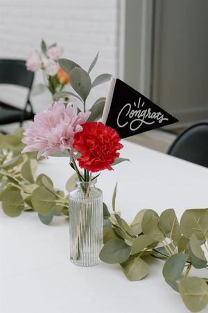 Glass vase with pink and red carnations, eucalyptus garland, and a black Congrats pennant flag on a white table.
