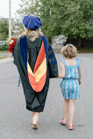 Graduate in blue cap and gown walks hand-in-hand with young girl on street, holding red bouquet.