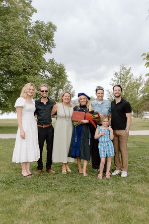 Group of seven people posing outdoors at a graduation celebration with a graduate in blue cap and gown holding diploma.
