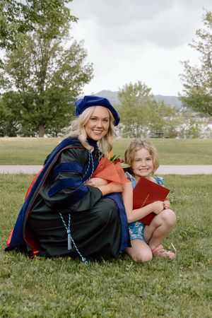 Woman in doctoral graduation regalia crouches beside young girl holding red diploma on green lawn.