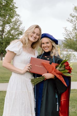 Two smiling women pose outdoors, one in a white dress and one in blue doctoral graduation regalia holding red roses.