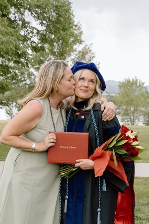 Woman kisses graduate in blue cap and gown holding University of Denver diploma and red roses.