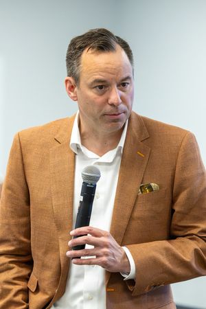 Man in tan blazer holding microphone speaking at indoor professional event or conference.