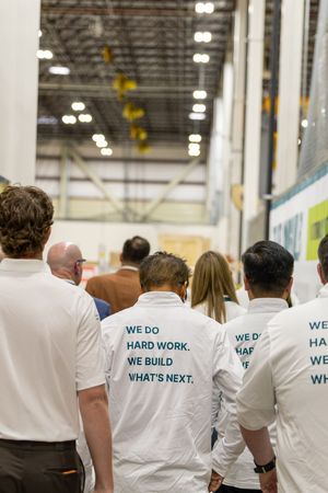 Group of workers in white t-shirts reading We Do Hard Work We Build Whats Next walking through industrial warehouse.