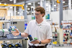 A young male engineer in safety glasses examines electronic components on a factory floor.