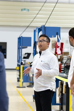 Man in white jacket gestures while speaking to colleagues in a modern industrial facility.