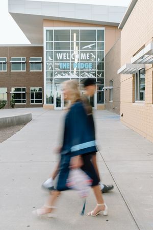 Two blurred graduates walk past a brick building with a Welcome to The Ridge sign in the background.