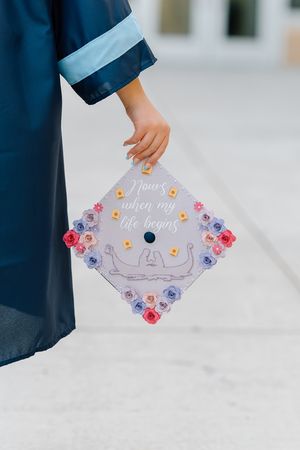 Close-up of graduation cap decorated with paper flowers and 'Now's when my life begins' quote.