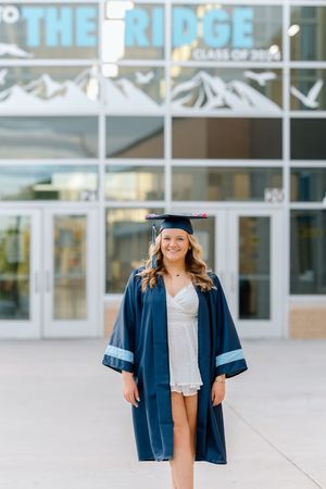 Smiling graduate in navy cap and gown poses outside The Ridge school building on a sunny day.
