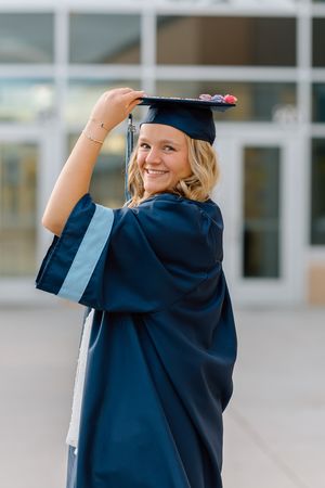 Smiling graduate in navy cap and gown holds mortarboard outdoors in front of a modern building.