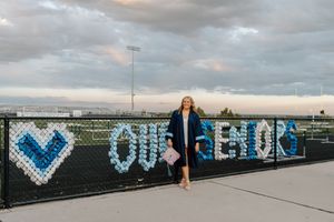 Woman in graduation gown poses by a fence decorated with blue and white flowers spelling OUHS outdoors.