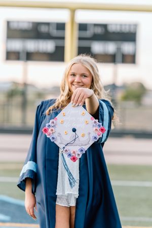 Smiling blonde graduate in navy gown holding decorated floral mortarboard cap toward camera.