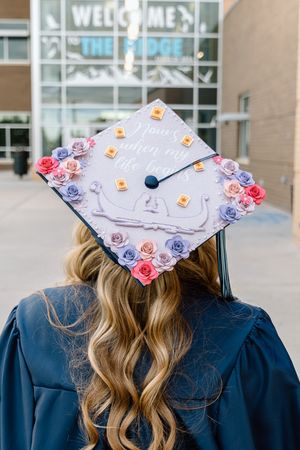 Graduate in navy gown showing decorated mortarboard cap with floral border and Tangled quote.