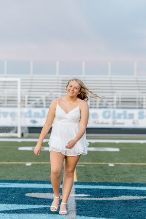 Smiling blonde graduate in white dress posing on blue turf football field at sunset.