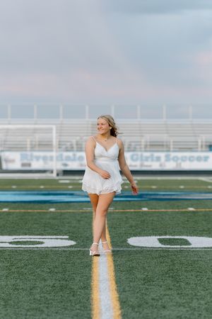 Young woman in white dress walking on soccer field at Riverdale Girls Soccer stadium during golden hour.