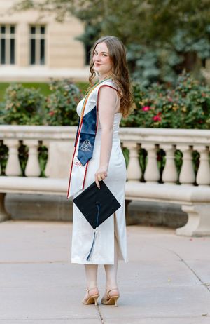 Young woman in white dress holding graduation cap with academic sash outdoors at university.